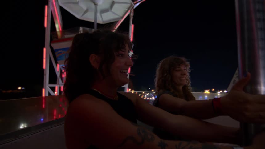 Two happy female friends smile and laugh together while riding a brightly lit Ferris wheel at a nighttime carnival