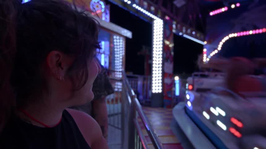 Close up of a young woman smiling as she watches a brightly lit, spinning ride at a funfair at night