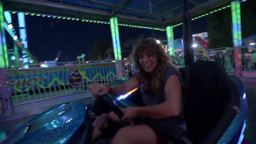 POV shot of a joyful woman laughing while driving a bumper car at a vibrant carnival at night