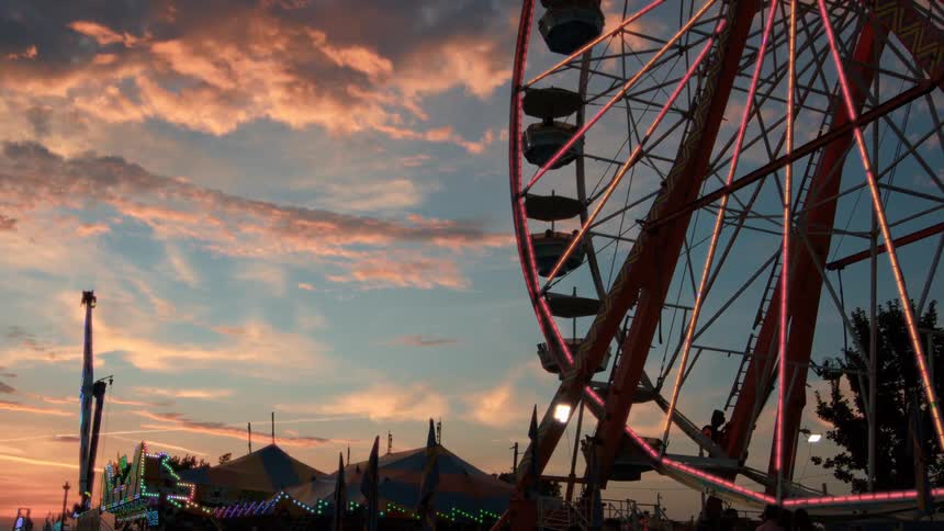 A brightly lit Ferris wheel and other carnival rides operate against a stunning sunset sky