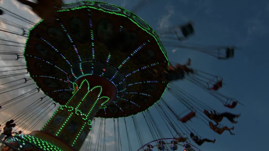 An exciting low-angle shot captures a brightly lit swing ride spinning people around at a carnival against the dusk sky