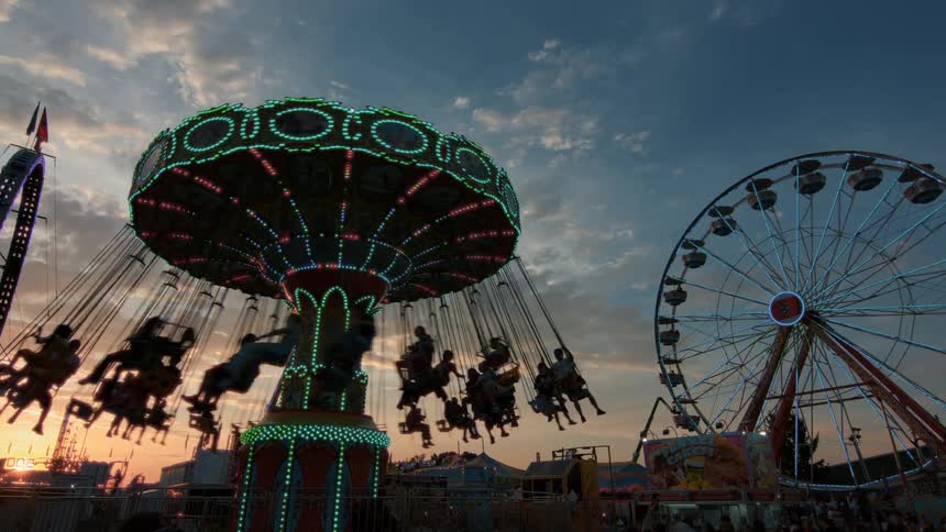Silhouetted people enjoy a spinning swing ride at a carnival with a brightly lit Ferris wheel in the background against a beautiful sunset sky