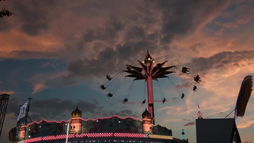 A brightly lit swing ride at a carnival spins passengers high into the air against a dramatic sunset sky
