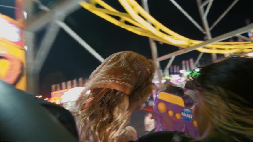 Two young women enjoy a thrilling roller coaster ride from a first-person perspective at a vibrant amusement park at night