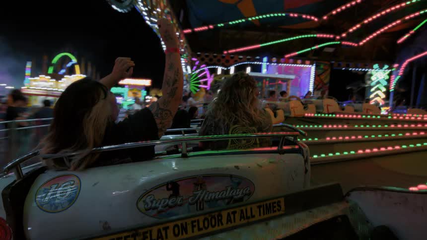 Two friends with their hands in the air enjoy a fast-spinning carnival ride surrounded by flashing lights at night