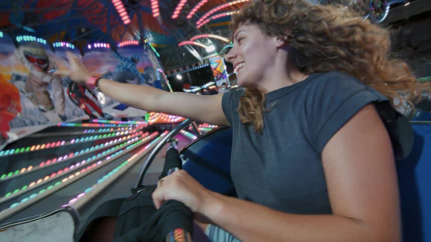 A joyful young woman with curly hair smiles while riding a fast, spinning amusement park ride at night, surrounded by colorful flashing lights