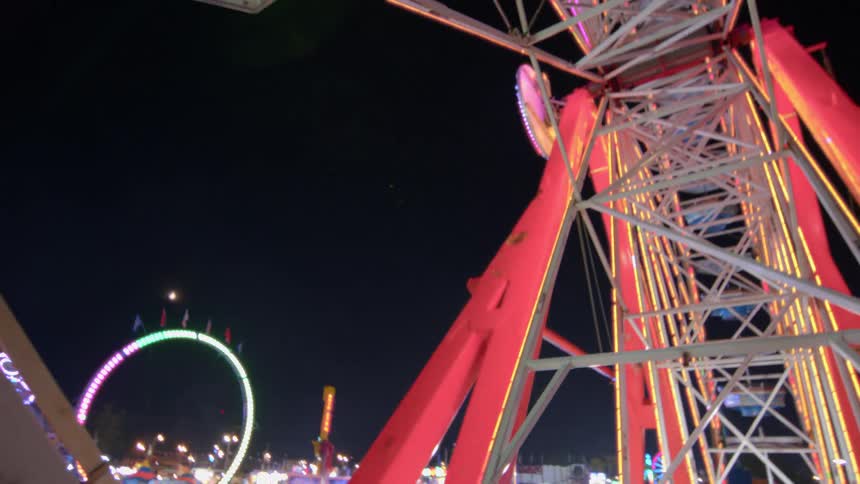 A low-angle view from a ride tilting up towards a giant, brightly lit Ferris wheel at a bustling carnival at night