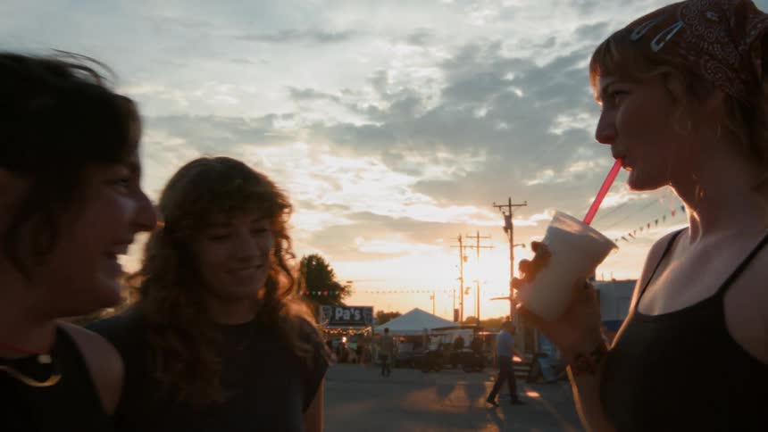Three happy friends laugh and chat while enjoying a milkshake at a fun outdoor fair during a beautiful sunset