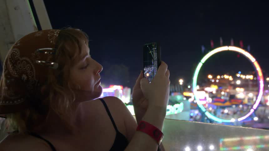 A woman in a bandana takes pictures on her phone from a Ferris wheel overlooking a brightly lit carnival at night in slow motion