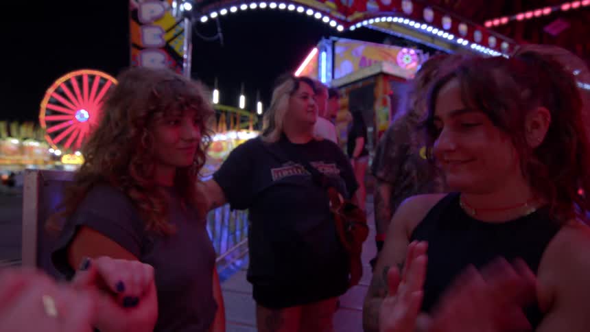A group of young women friends hold hands and laugh while waiting for a carnival ride at night