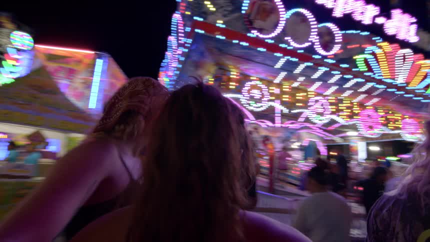 Two young women embrace and smile while on a fast-spinning carnival ride surrounded by the vibrant, colorful lights of the fair at night
