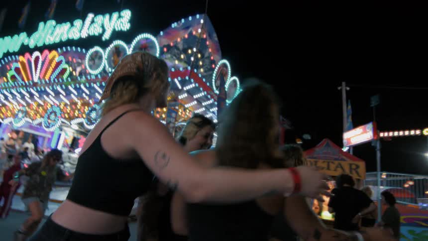 A group of young women laugh and dance together while enjoying the bright lights and thrilling rides at a carnival at night