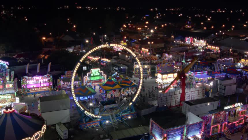 A vibrant carnival glows at night as a brightly lit circular ride spins, seen from a high-angle aerial view