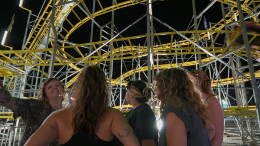 A group of young women have an exciting conversation while watching a brightly lit roller coaster at a carnival at night
