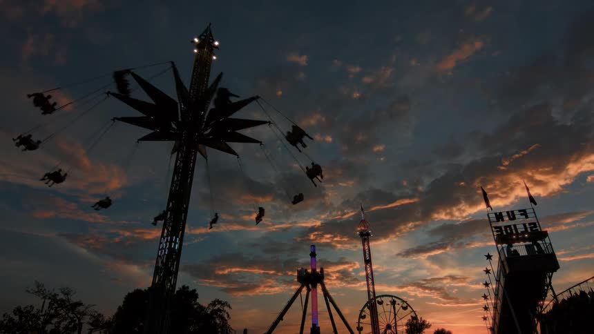 A tall, illuminated swing ride spins silhouetted people high into the air against a vibrant sunset sky at a carnival