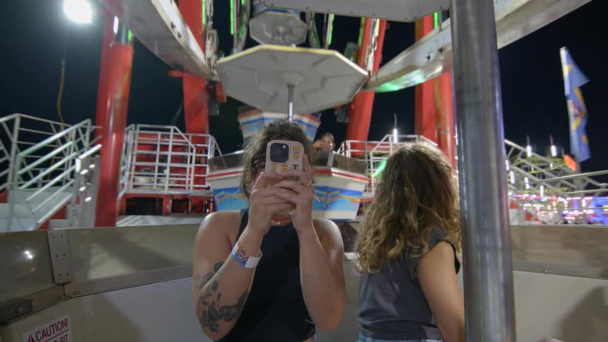 Two happy young women laugh and use a smartphone to capture the view from a spinning Ferris wheel at a carnival at night