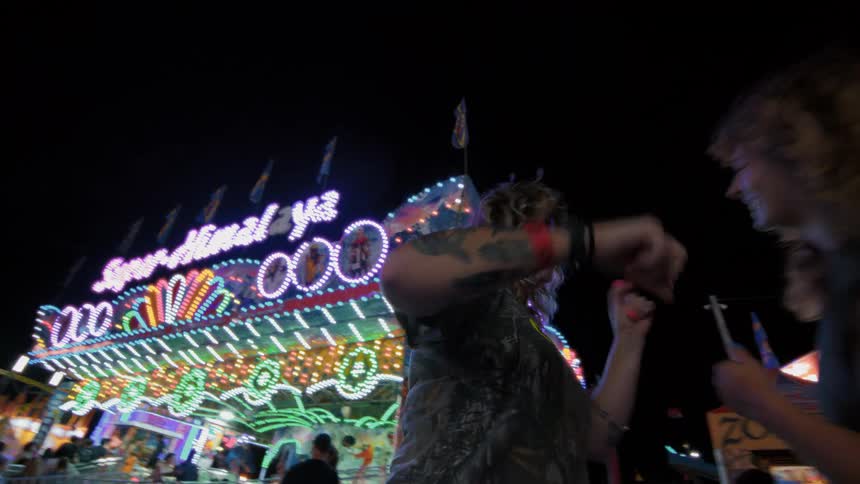 Three energetic women dance and laugh together in slow motion in front of a colorful, brightly lit ride at a carnival at night