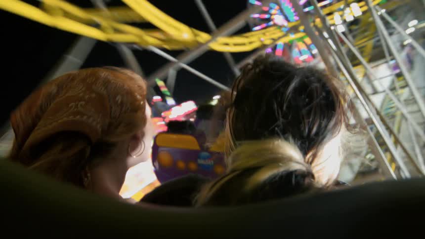 From a back seat perspective, two friends enjoy a thrilling roller coaster ride through a vibrant carnival at night