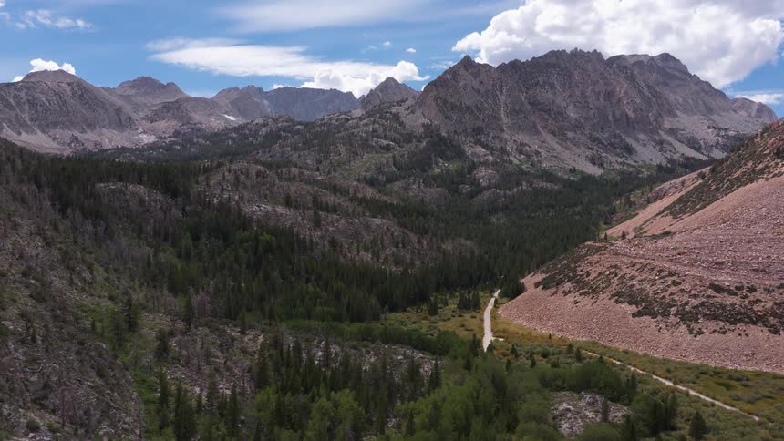 A slow aerial push forward reveals a beautiful, rugged mountain valley with a winding dirt road and green forest under a partly cloudy sky