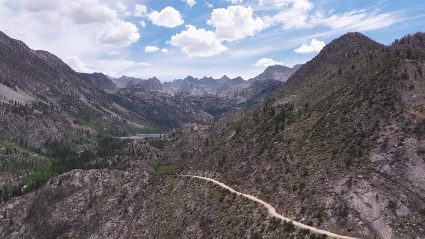 A majestic aerial pullback shot reveals a vast, rugged mountain valley with a winding dirt road leading towards a distant alpine lake