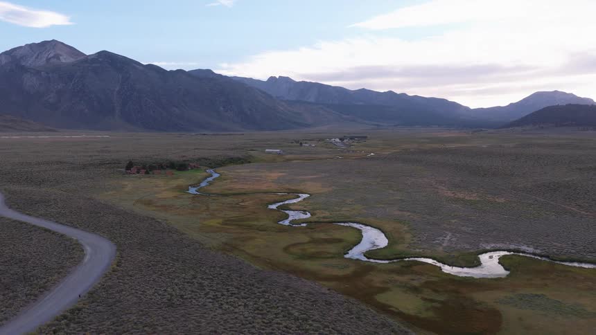 A majestic aerial view reveals a remote valley with a winding river and a dirt road nestled at the base of a mountain range at dusk