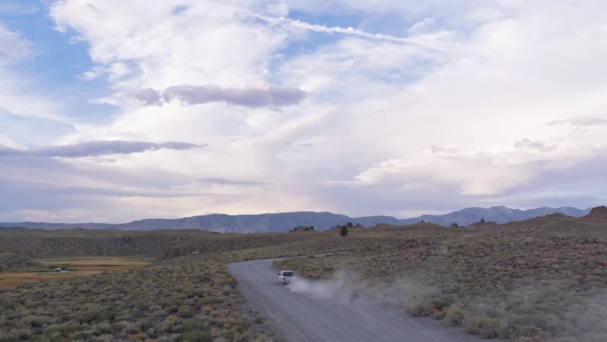 An aerial view follows a white pickup truck as it kicks up dust while driving down a remote gravel road through a vast, mountainous landscape at twilight