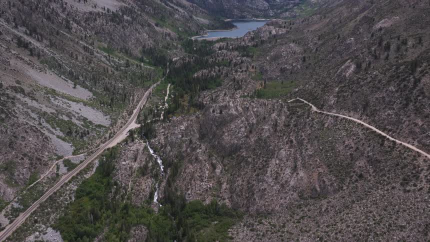 A dramatic aerial view tilts up from a cascading waterfall to reveal a vast, rugged mountain valley with a pristine alpine lake