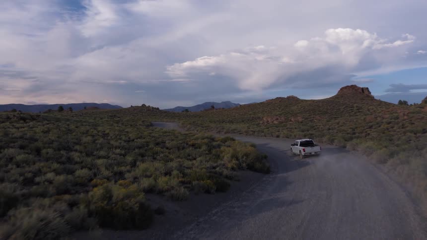 An aerial view follows a silver truck as it kicks up dust driving on a winding dirt road through the remote, arid wilderness at dusk