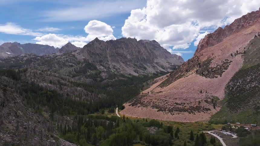 Expansive aerial view pulling back over a scenic mountain valley, showcasing a dense forest, winding trail, and rugged peaks