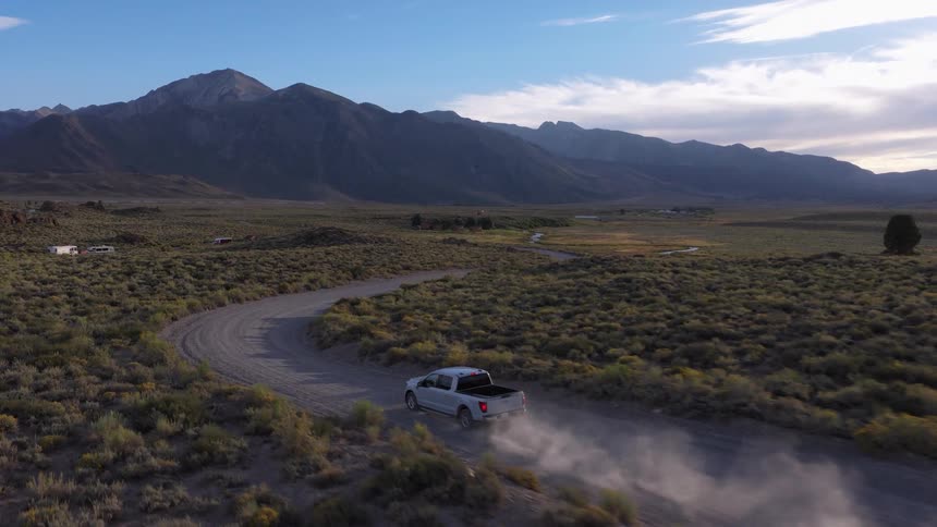 An aerial view follows a silver pickup truck driving along a dusty, winding road through a scenic mountain valley at sunrise