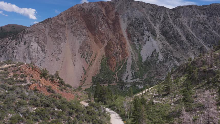 An aerial view pushes forward over a dirt road towards a massive, colorful mountain with exposed rock faces under a blue sky