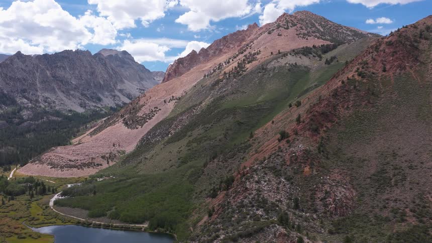 An aerial shot pulls back from a dramatic mountain peak to reveal a serene alpine lake and verdant valley below
