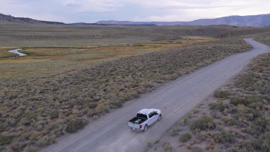 An aerial view follows a white pickup truck driving along a dusty, winding dirt road through a vast, remote sagebrush landscape at dusk