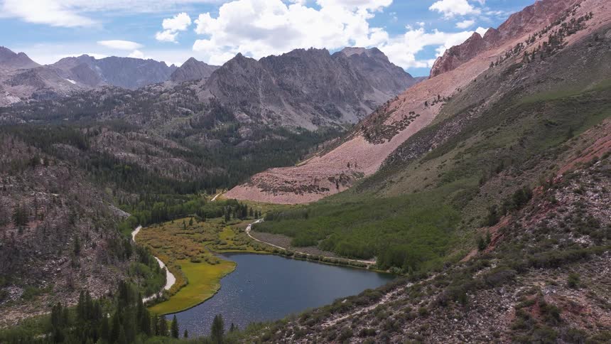 A stunning aerial view pulls back to reveal a serene alpine lake nestled in a vast mountain valley with lush forests and rugged peaks