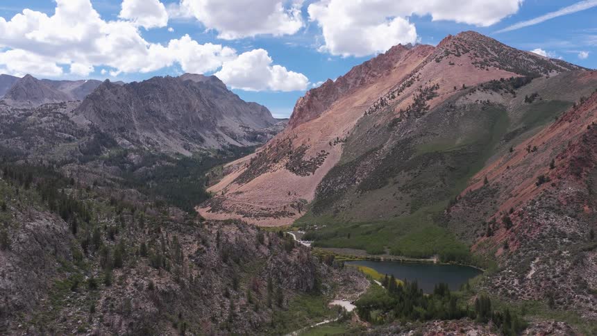 A stunning aerial view reveals a pristine alpine lake nestled in a rugged mountain valley under a partly cloudy sky