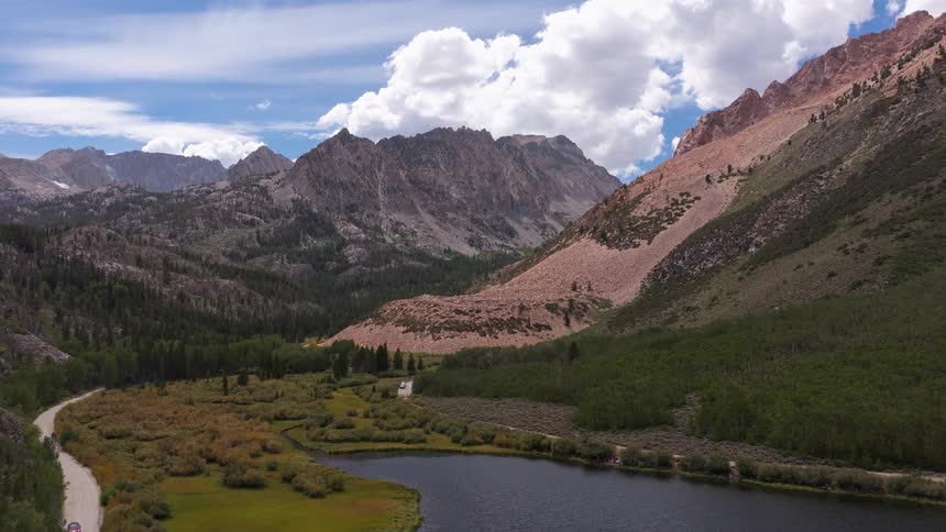 An epic aerial reveal of a stunning mountain lake surrounded by rugged peaks, lush forests, and a winding dirt road on a beautiful summer day