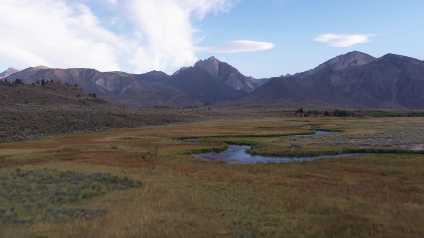 A stunning aerial view ascends over a tranquil river winding through a vast meadow towards a majestic mountain range at dusk