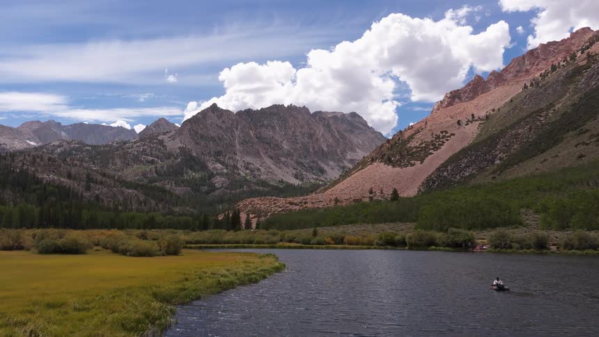 An aerial view slowly advances over a calm mountain lake where people are boating, revealing a majestic and rugged mountain landscape