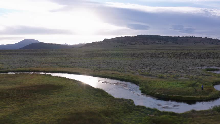 A sweeping aerial view reveals a lone fly fisherman casting his line in a tranquil, winding river that meanders through a vast mountain meadow at sunrise
