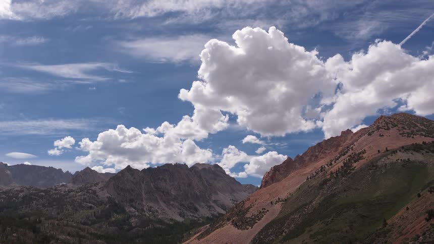 A slow zoom out from a bright, cloudy sky reveals a majestic mountain valley with a pristine alpine lake