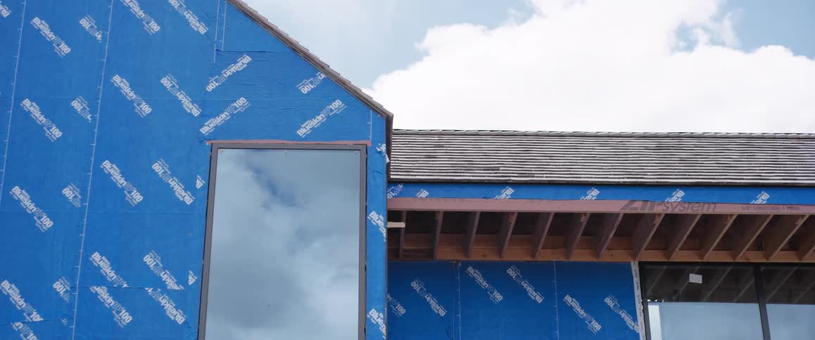 The exterior of a modern home under construction, showing the blue weather barrier, roof, and a large window reflecting the cloudy sky