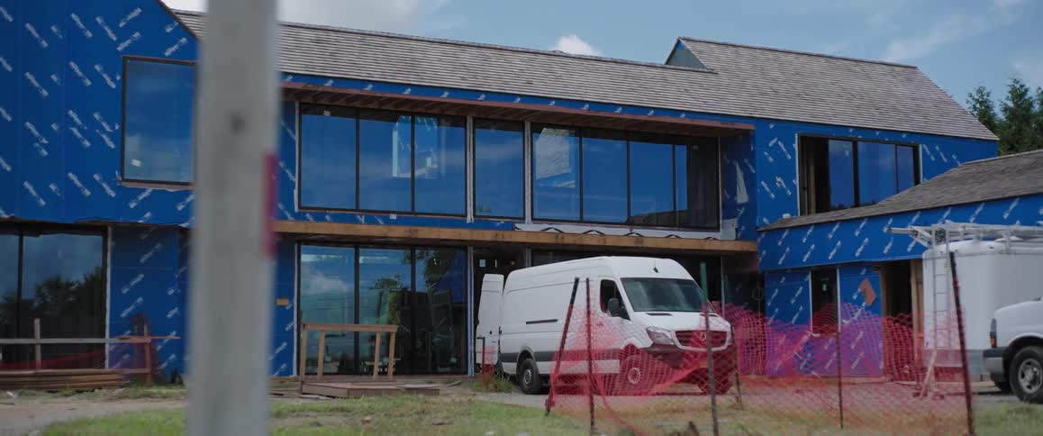 A modern two-story house under construction with blue weatherproofing wrap and a white work van parked outside
