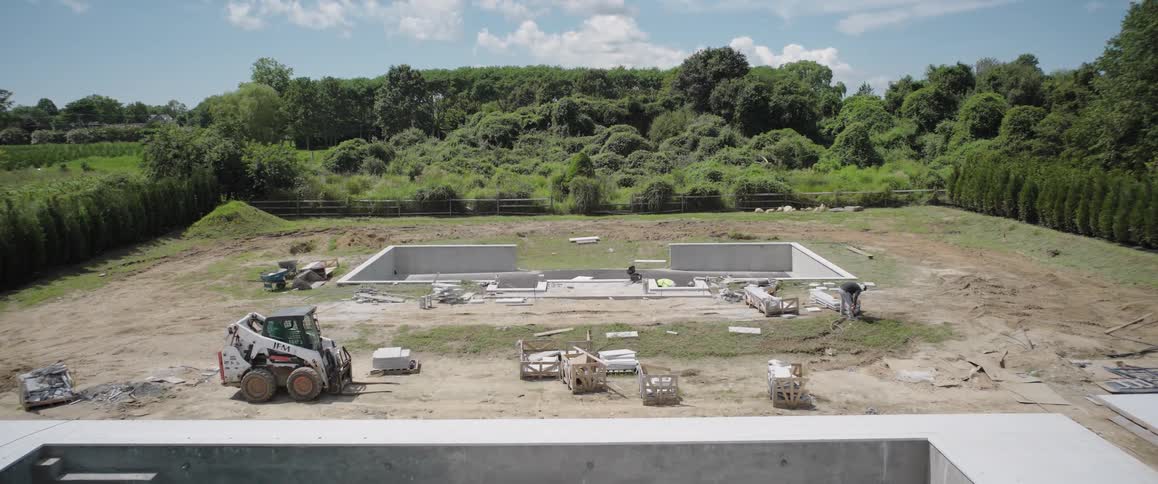 Wide static shot of a large in-ground swimming pool under construction in a spacious backyard on a sunny day