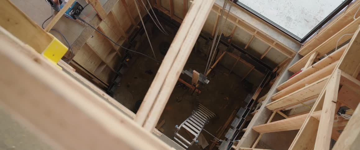 A high-angle, slow-moving view down an unfinished elevator shaft inside a wood-framed building under construction