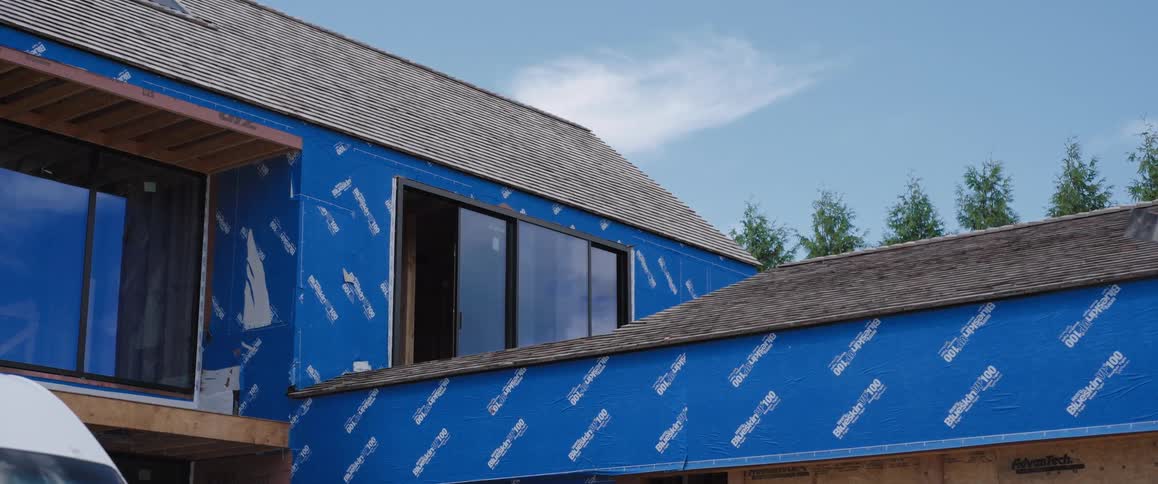 A new home under construction with blue house wrap covering the exterior walls and a newly shingled roof under a blue sky