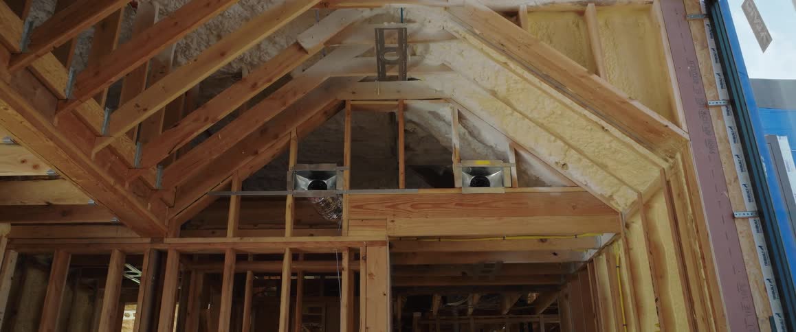 A downward pan of the wooden frame of a house under construction shows the spray foam insulation and HVAC vents in the ceiling