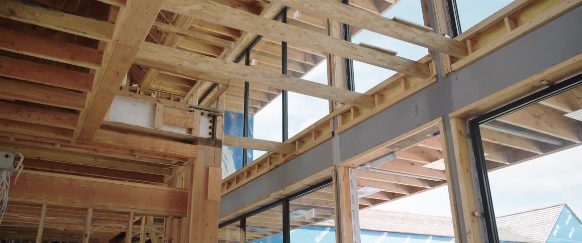 Camera pans across the wooden frame of a modern house under construction, revealing exposed beams, joists, and large windows