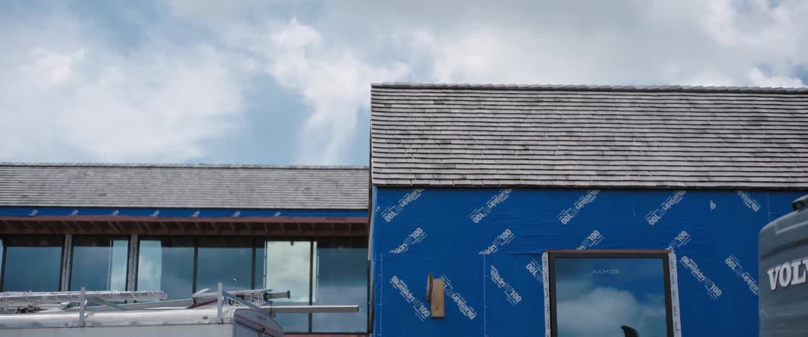 A slow rightward pan reveals a modern home under construction, its exterior wrapped in a blue vapor barrier beneath a newly shingled roof