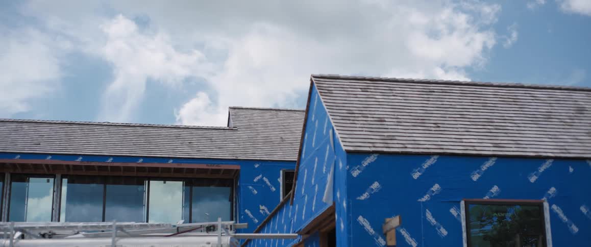 Slow zoom out time-lapse of clouds passing over a modern house under construction with blue weather-resistant barrier