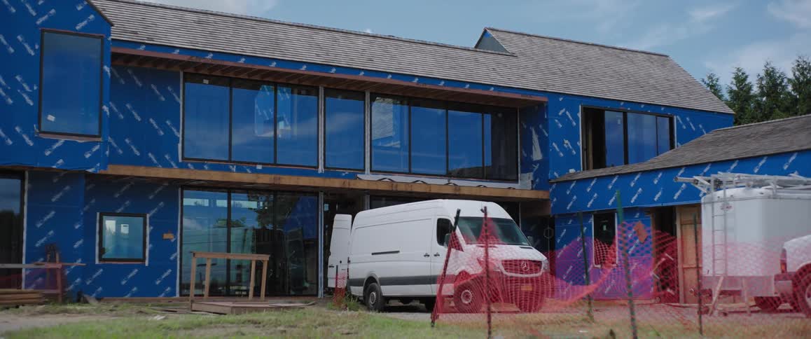 A white work van is parked at a modern house construction site with blue exterior sheathing during the day